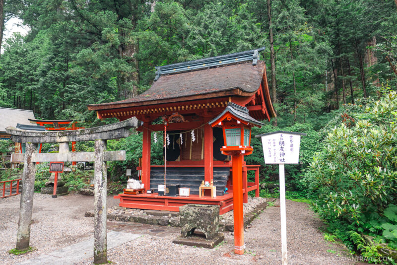 A small red Shinto shrine with a traditional gate stands surrounded by lush green trees at Nikko Futarasan Jinja. Offerings are placed on the steps, and a white sign with Japanese text is in front of the shrine.