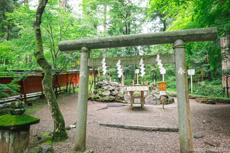 A stone torii gate stands at the entrance of Nikko Futarasan Jinja, surrounded by lush green trees, with sacred white paper streamers hanging from the gate and a small shrine structure in the background.