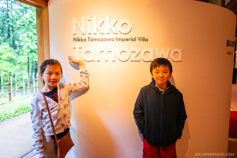 Two children stand inside near a curved wall sign that reads Nikko Tamozawa Imperial Villa Memorial Park. One girl points to the sign, while a boy stands beside her. Green trees are visible through the window on the left.
