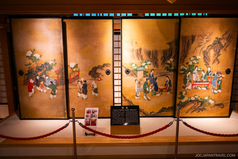 Four traditional Japanese folding screens with detailed paintings of people in historical clothing, displayed in a museum setting at Nikko Tamozawa Imperial Villa Memorial Park, stand behind red ropes with an information plaque in front.