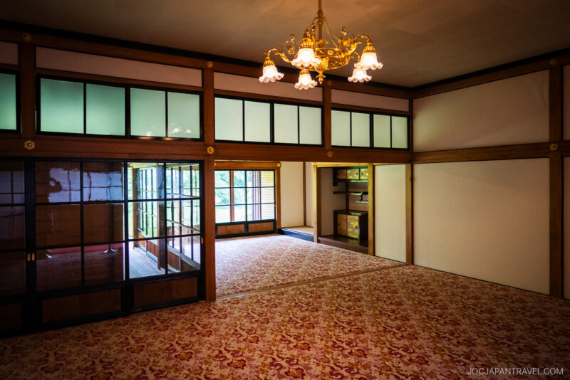 A spacious traditional Japanese room with tatami mats, wood details, and a vintage chandelier, reminiscent of Nikko Tamozawa Imperial Villa Memorial Park, overlooking a garden through large sliding glass doors.