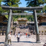 Nikko Toshogu Shrine (日光東照宮) – Japan’s Most Ornate Shinto Shrine Visitors walk through a large torii gate toward ornate temple buildings at Nikko Toshogu Shrine, surrounded by tall trees at this historic site in Japan, with steps leading up to the main shrine.