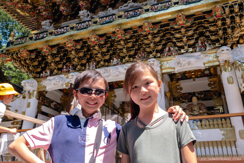 Two smiling children pose together in front of the ornate Nikko Toshogu Shrine, its gold decorations and intricate carvings gleaming in the sunlight. Visitors can be seen exploring the background.