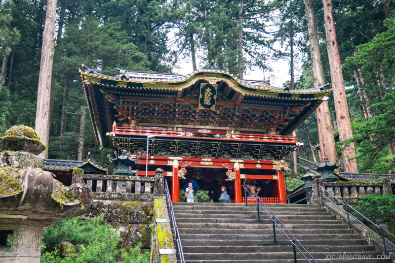 A colorful traditional Japanese temple, Nikkosan Rinnoji Taiyuin, with intricate carvings and gold details stands among tall trees. Several visitors are on the stone steps leading up to the entrance.