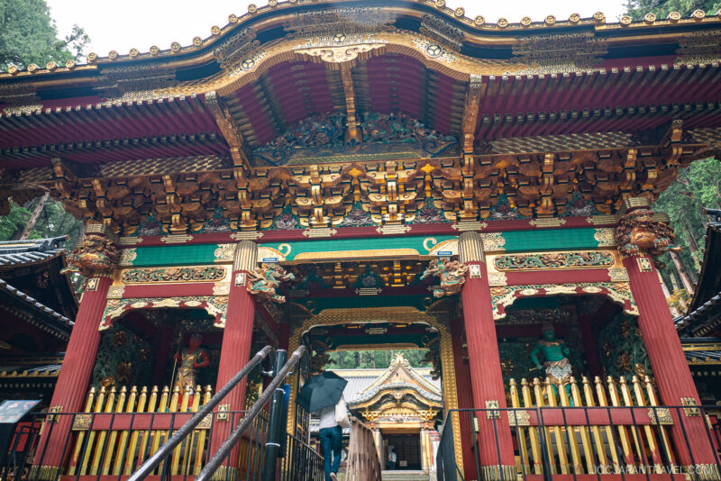 Large, ornate traditional Japanese temple gate at Nikkosan Rinnoji Taiyuin with intricate gold, red, and green carvings, wooden beams, and statues, surrounded by trees and a visitor ascending the stairs.