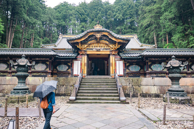 A person with an umbrella walks toward Nikkosan Rinnoji Taiyuin, an ornate Japanese temple surrounded by tall trees. Stone lanterns flank the steps leading up to the temple’s richly decorated entrance.