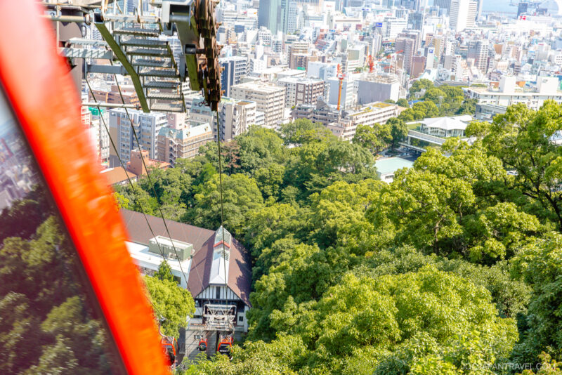 Aerial view from a red cable car on the Nunobiki Ropeway descending toward a station surrounded by green trees and the Herb Gardens, with a cityscape of tall buildings and streets in the background.