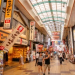 A busy indoor shopping street in Namba, Osaka, with colorful signs in Japanese, shops on both sides, and people walking along the tiled walkway under a glass roof.