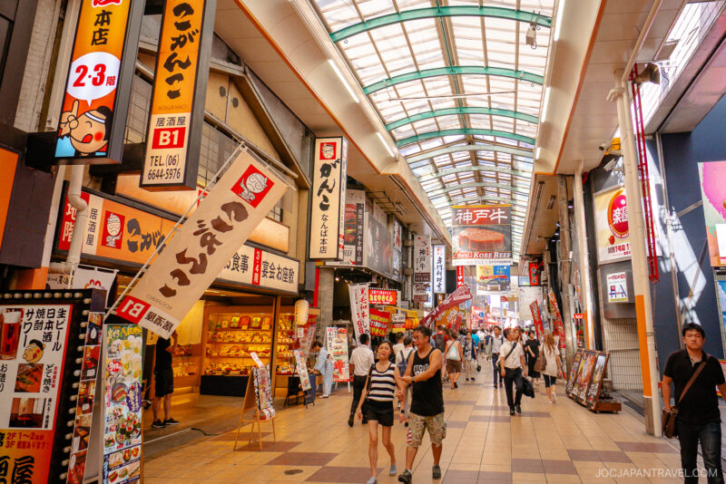 A busy indoor shopping street in Namba, Osaka, with colorful signs in Japanese, shops on both sides, and people walking along the tiled walkway under a glass roof.