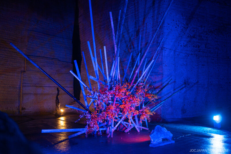 A modern art installation at the Oya History Museum features intersecting metal rods in a starburst pattern, adorned with bright red flowers and illuminated by blue and purple lighting in a dark, spacious room with textured walls.