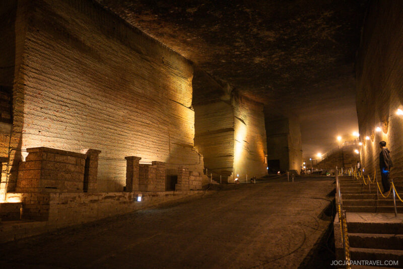 An underground stone quarry with tall, textured walls lit by warm lights. At the Oya History Museum, a person stands on the right near a staircase with a rope barrier, and the vast space extends into the distance.