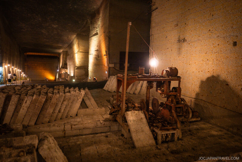 An underground stone quarry with tall, illuminated walls, old rusty mining machinery, and rows of large stone blocks—the Oya History Museum’s dim lighting creates deep shadows and a mysterious atmosphere.