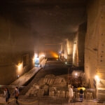 Oya History Museum (大谷資料館) – An Underground World of Stone and Art A large, illuminated underground stone quarry with high ceilings and massive walls, the Oya History Museum features pathways bordered by ropes. Several people walk above, observing construction materials and equipment displayed on the floor below.