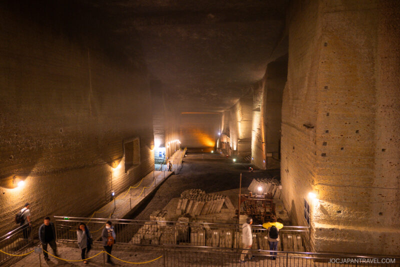 A large, illuminated underground stone quarry with high ceilings and massive walls, the Oya History Museum features pathways bordered by ropes. Several people walk above, observing construction materials and equipment displayed on the floor below.