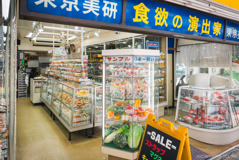 A shop in Japan displays realistic Plastic Food Sampuru models in glass cases, with colorful dishes like sushi and desserts. A yellow sale sign stands outside, and Japanese signage is visible above the entrance.