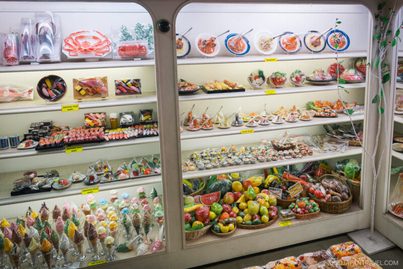 A display case filled with realistic Plastic Food Sampuru replicas, including sushi, noodles, fruit, desserts, and drinks—commonly showcased in Japanese restaurant windows to illustrate menu items.