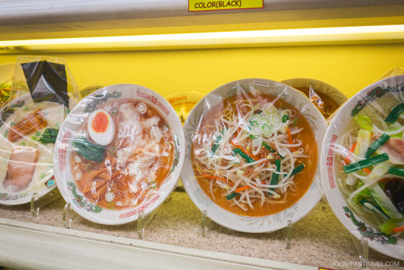 Plastic Food Sampuru display bowls of ramen with various toppings, including boiled eggs, sliced pork, bean sprouts, and green onions, are lined up on a shelf in a brightly lit setting.