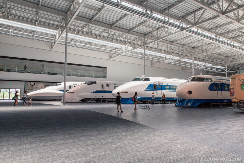 Several people observe a display of four different high-speed Shinkansen train models inside the spacious, modern SCMaglev and Railway Park exhibit hall with large windows and high ceilings.