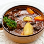 A brown ceramic bowl filled with Seiyoken beef stew, featuring chunks of meat, potatoes, carrots, broccoli, and a pearl onion in a rich, dark broth, placed on a white lace doily.