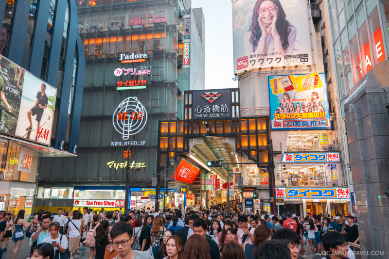 A bustling street scene in Osaka Shinsaibashi, Japan, filled with crowds of people and surrounded by colorful storefronts, bright neon signs, and billboards in Japanese.