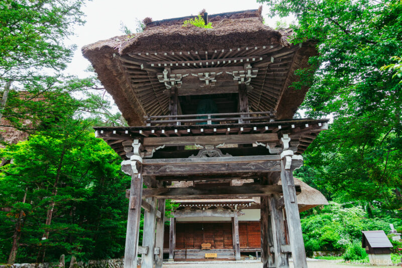 A traditional wooden Japanese gate with a thatched roof stands surrounded by lush green trees in Shirakawa-go, leading to a building in the background. The structure shows weathered wood and detailed craftsmanship.