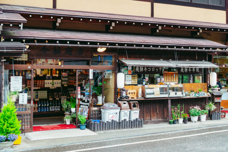 Traditional Japanese storefront with wooden architecture reminiscent of Shirakawa-go, shelves of bottles visible inside, food items and plants displayed outside, and lanterns hanging by the entrance on a rainy street.