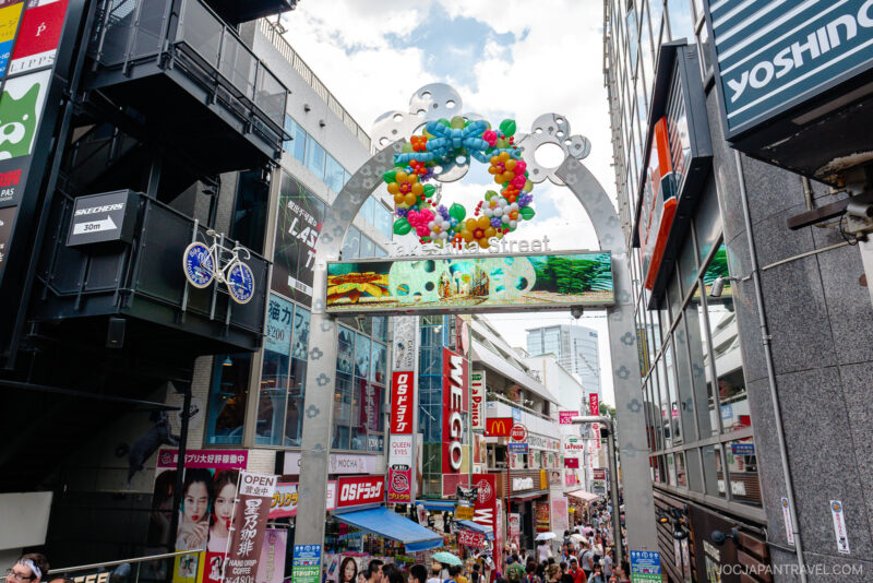 A bustling Takeshita Street Harajuku scene in Tokyo, Japan, filled with colorful shops, signs, and a decorative entrance arch with balloons. Crowds of people stroll beneath the arch on a sunny day.