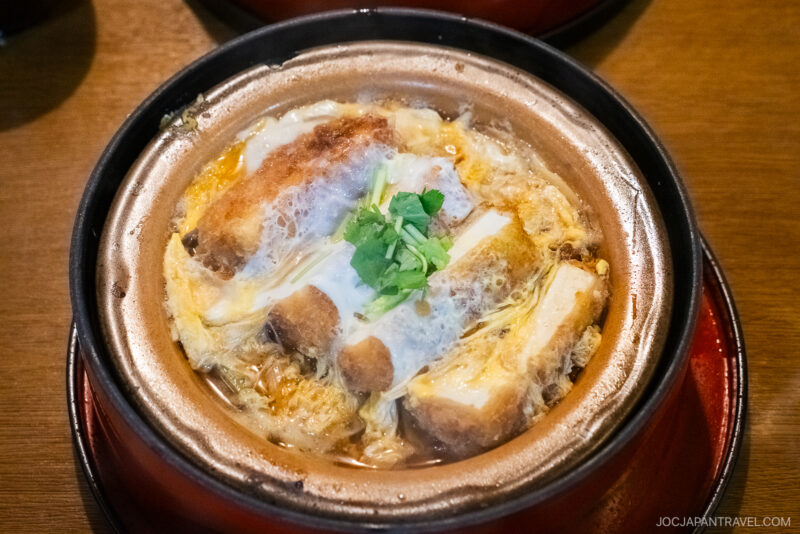 A bowl of katsudon from Tamura Ginkatsu-tei, featuring breaded pork cutlet, onions, and egg cooked in savory broth, garnished with chopped green onions, served in a rustic clay pot on a wooden table.