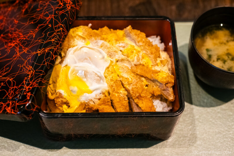 At Tamura Ginkatsu-tei, enjoy a lacquered bento box with breaded pork cutlet topped with egg over rice, partially covered by a decorative lid, and served with miso soup on a green placemat.