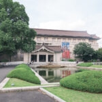 Tokyo National Museum stands behind a pond and manicured bushes, with a few people walking and sitting on benches nearby. The sky is partly cloudy.