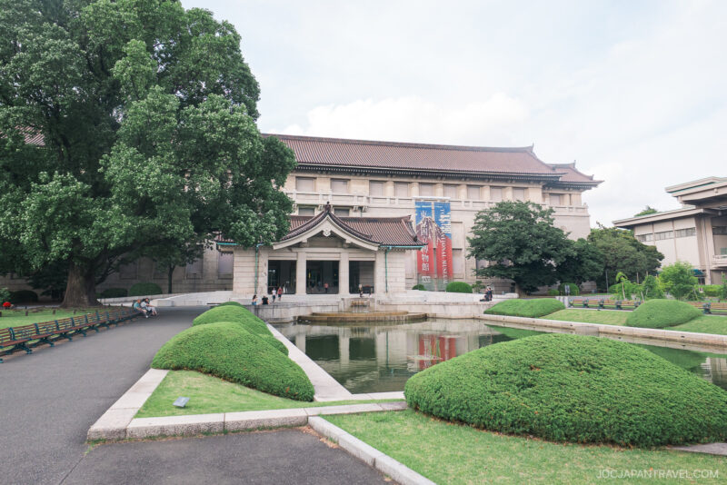 Tokyo National Museum stands behind a pond and manicured bushes, with a few people walking and sitting on benches nearby. The sky is partly cloudy.