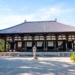 Toshodai-ji (唐招提寺): Nara’s Temple of Peace The traditional Japanese wooden temple building of Nara Toshodai-ji, with its large tiled roof, stands surrounded by greenery under a clear blue sky. A stone lantern is centered in front on the gravel courtyard.