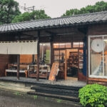 Touki Yufuin is a traditional Japanese pottery shop with a tiled roof, wooden exterior, large windows, and pottery displayed on a table outside. Its rainy, and lush greenery surrounds the building.