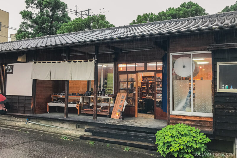 Touki Yufuin is a traditional Japanese pottery shop with a tiled roof, wooden exterior, large windows, and pottery displayed on a table outside. Its rainy, and lush greenery surrounds the building.