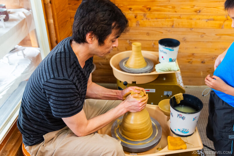 A man shapes clay on a pottery wheel inside a wooden room at Touki Yufuin, while a child stands nearby watching. Buckets and pottery tools are placed on the table, completing the serene scene.