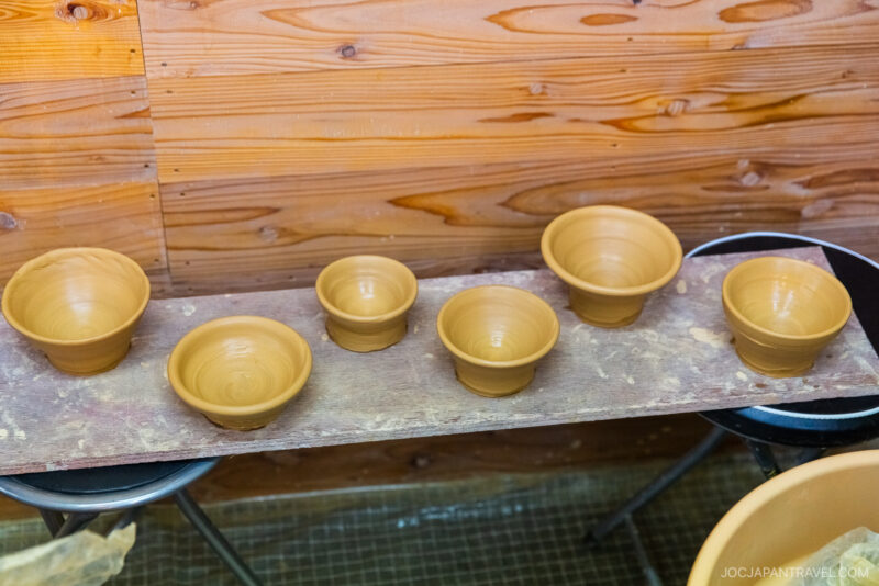 Six unfinished Touki Yufuin clay bowls of varying sizes are arranged on a wooden plank inside a pottery workshop with wooden walls in the background.
