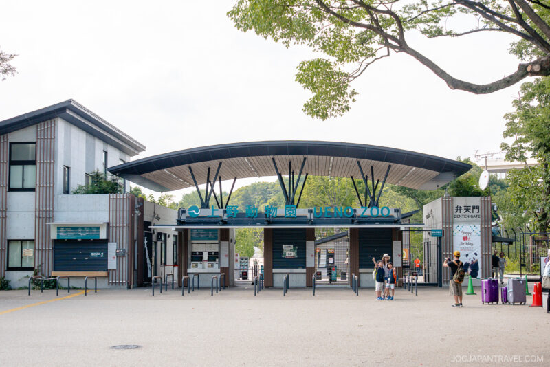 The entrance gate of Ueno Zoo in Tokyo, Japan, with people standing near ticket counters under a curved roof. Surrounding buildings and trees frame the lively Ueno Zoo area on a clear day.