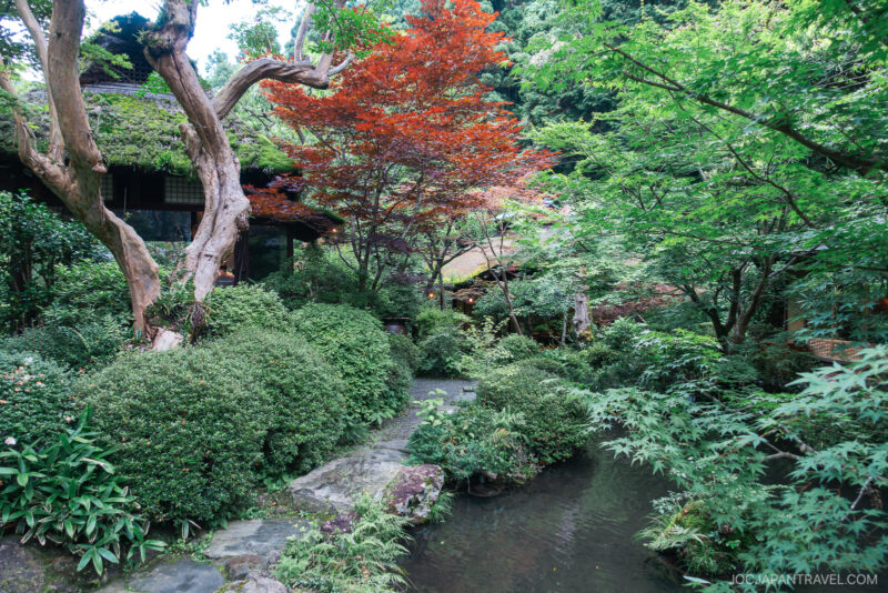Serene Japanese garden with a small stream, lush green foliage, a red maple tree, stone path, and traditional wooden buildings partially hidden by trees—evoking the tranquil ambiance of Ukai Toriyama.