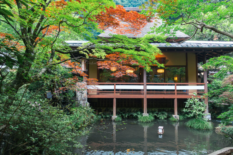 A serene Japanese garden with vibrant green and red maple trees surrounds Ukai Toriyama, a traditional wooden building overlooking a calm pond that reflects the lush foliage and a decorative lantern.