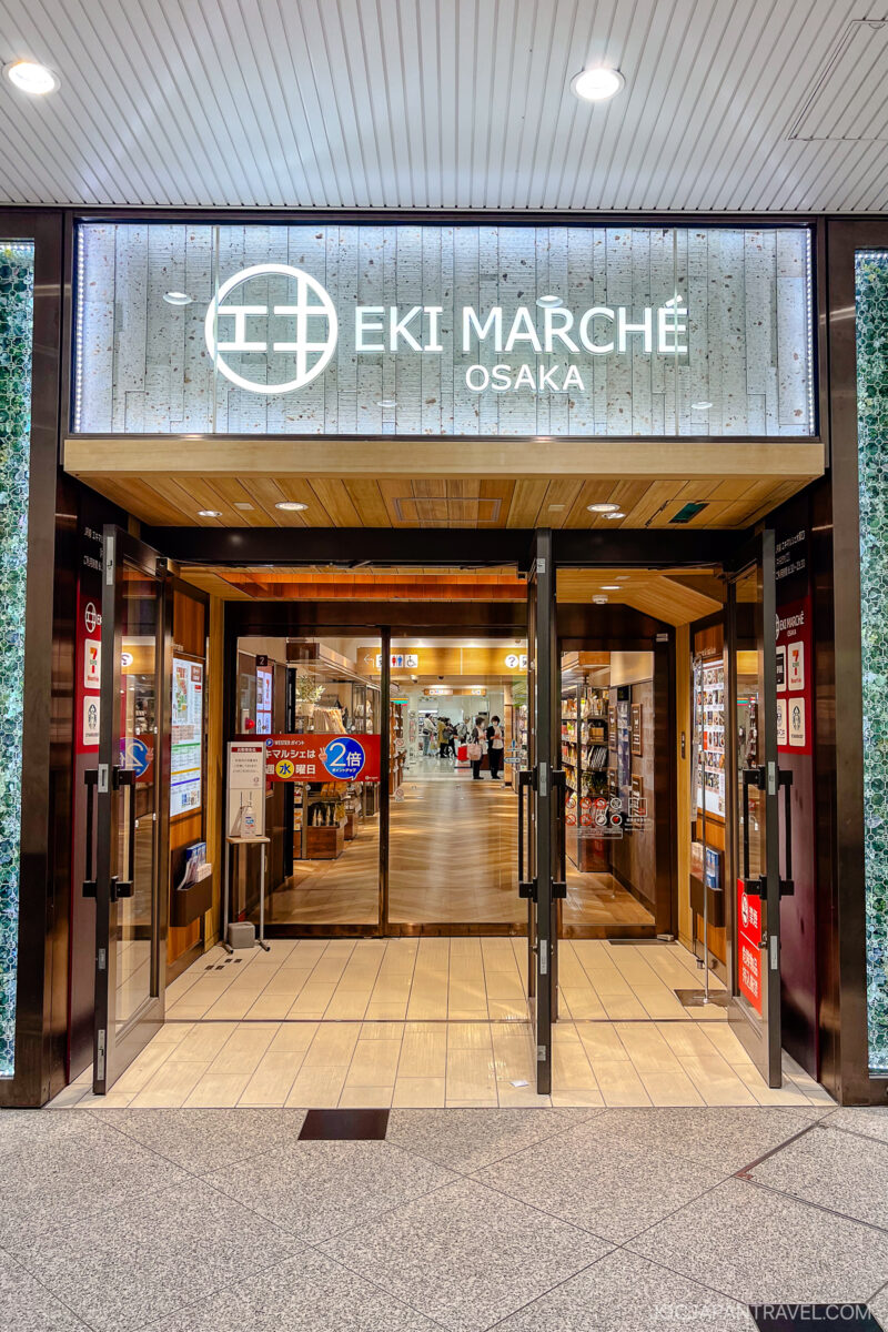 Entrance to Eki Marché Osaka, a shopping and dining area in Osaka Umeda. The glass doors are open, with signs visible at the entrance. The well-lit interior reveals shops and people inside.