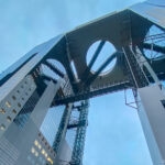 A dramatic upward view of the Umeda Sky Building in Osaka, Japan, showcases its two tall towers connected by a futuristic circular observation deck with escalators and bridges, all set against a cloudy sky.
