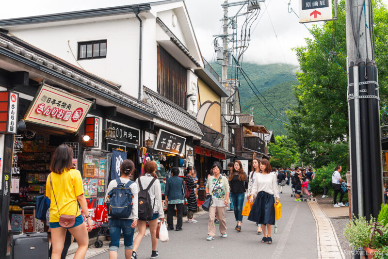 People walk along Yufuin Yunotsubo Kaido, a lively shopping street lined with traditional buildings, shops, and signs in Japanese. Trees and distant green hills are visible in the background, adding to the area’s vibrant atmosphere.
