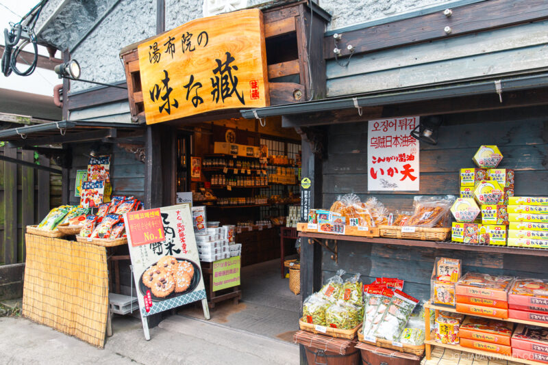 A traditional Japanese shop on Yufuin Yunotsubo Kaido with wooden walls, displaying snacks, packaged goods, and colorful signs in Japanese at the entrance. Inside, shelves are stocked with various products. The atmosphere is cozy and inviting.
