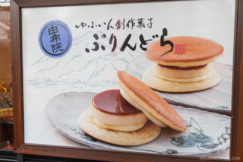A display shows two Japanese desserts: custard pudding sandwiched between two round, fluffy dorayaki pancakes, set against a backdrop featuring Yunotsubo Kaido mountain illustrations and Japanese text.