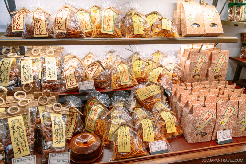 Bags and boxes of Japanese snacks neatly arranged on shelves, with handwritten yellow labels and price tags, inside a brightly lit shop on Yufuin Yunotsubo Kaido.