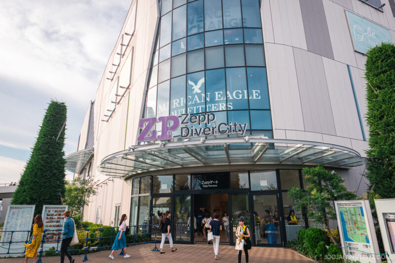 People walk past the entrance of Zepp DiverCity Odaiba, a modern multi-story building with large glass windows, American Eagle signage above, and greenery surrounding the entrance.