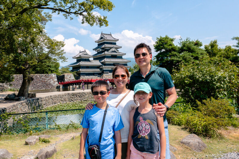 A smiling family of four poses outdoors in front of Matsumoto Castle, with a stone bridge, moat, and lush green trees beneath a bright blue sky.