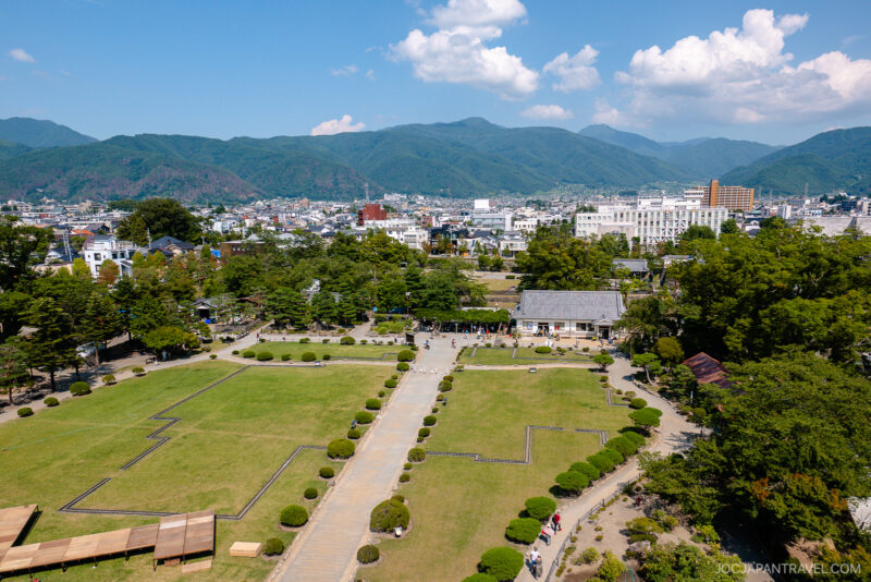 Aerial view of a park with trimmed lawns, pathways, and visitors, set against a cityscape featuring Matsumoto Castle and distant green mountains under a blue sky with scattered clouds.