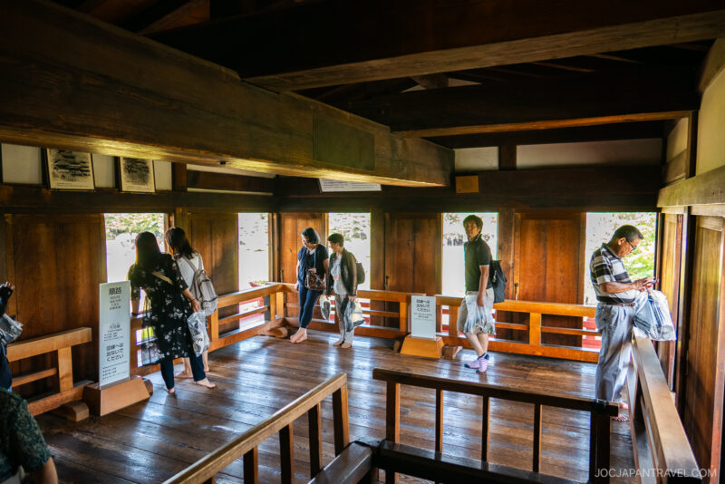 A group of people, some wearing masks, stand and walk inside a traditional wooden Japanese building at Matsumoto Castle, looking out the windows and reading information signs as sunlight filters through and illuminates the wooden interior.