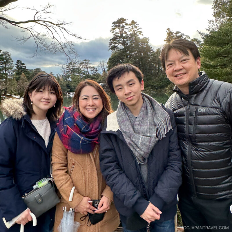 Four people stand close together outdoors, smiling at the camera. Bundled in winter jackets and scarves, they enjoy a chilly day in a park, perhaps sharing stories about Japanese culture amid the trees and cloudy sky.